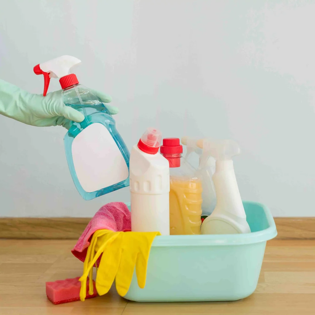 Office cleaning supplies in a bucket including sprays, gloves, cloths, and a mop on a floor 