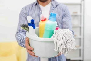 Man holding bucket with office cleaning products including sprays, mop, and brush in workspace