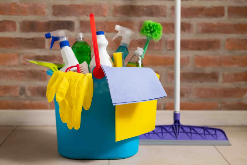 Office cleaning supplies in a bucket—sprays, gloves, cloths, and a mop—on a floor near a brick wall