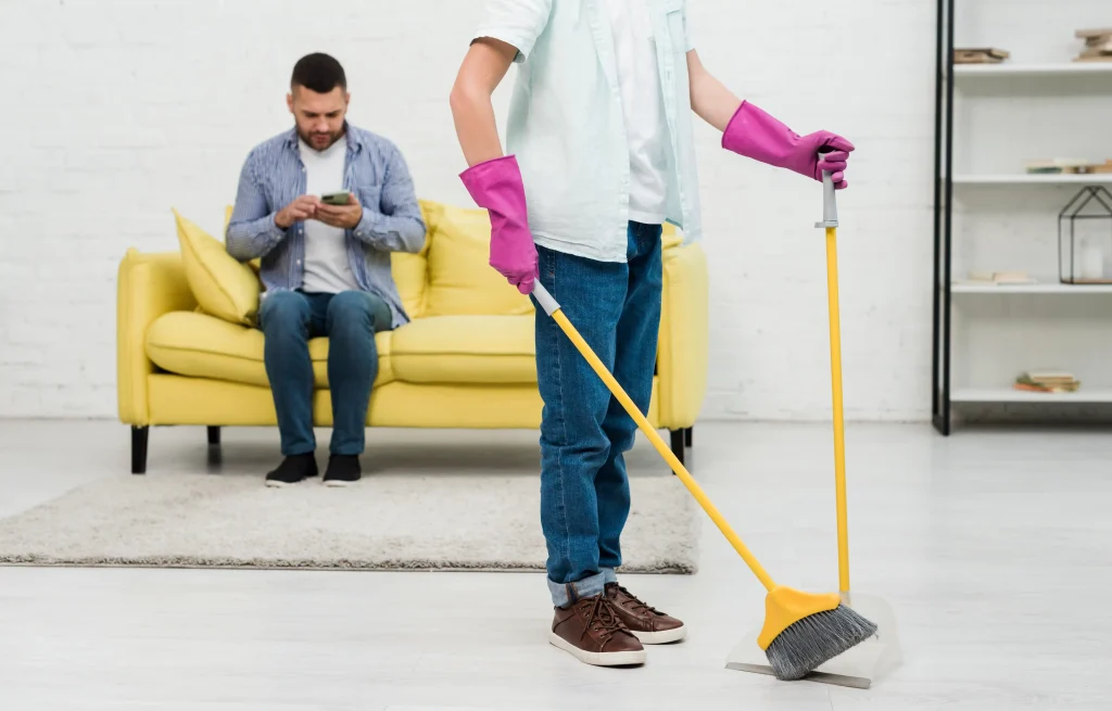 Person sweeping a bright living room floor while another person sits on a yellow sofa during move-out cleaning.
