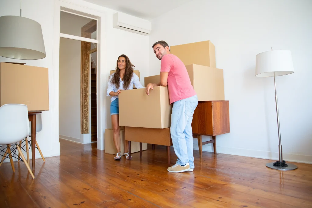Couple carrying moving boxes in a bright apartment during move-out cleaning with packed cardboard boxes and furniture.